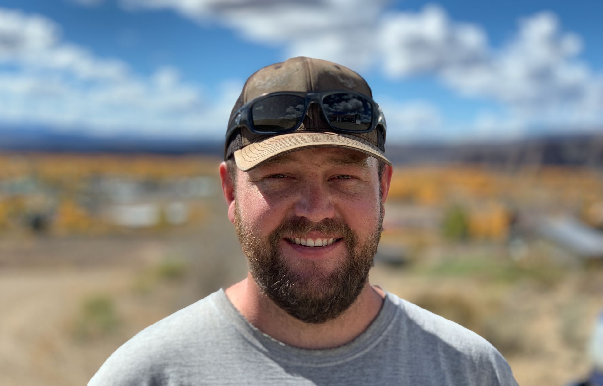 Smiling man wearing a cap and sunglasses on a sunny outdoor trail with mountains in the background