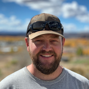 Smiling man wearing a cap and sunglasses, standing outdoors with a blurred landscape and blue sky behind him