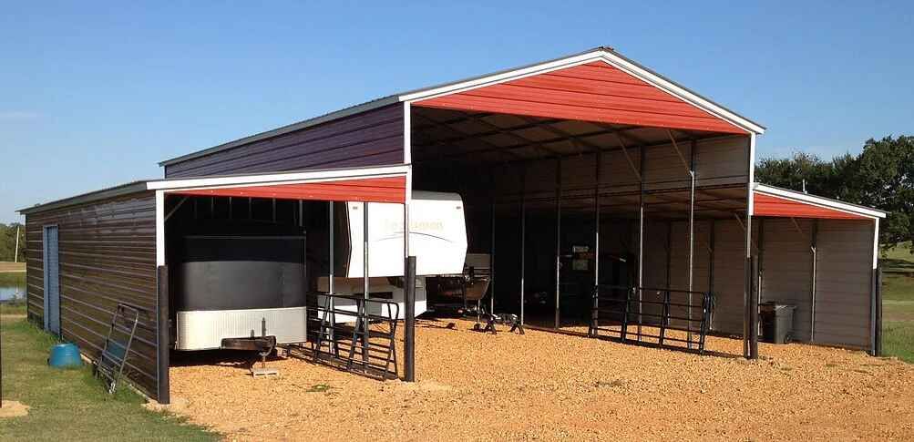 A large metal barn with a red roof, protecting a trailer and RV, under a clear blue sky.