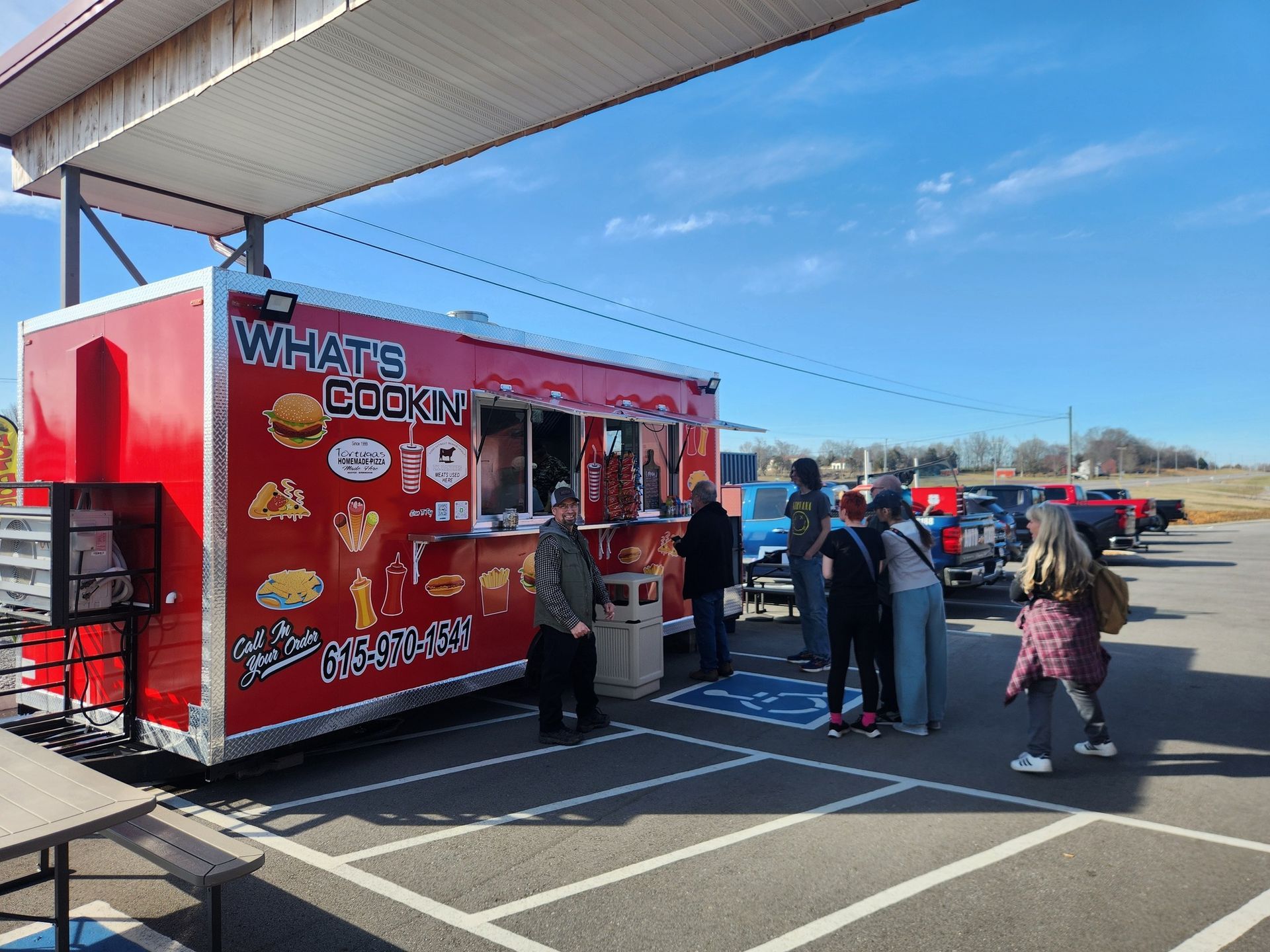 Red food truck "What's Cookin" with a line of people waiting. Sunny day, blue sky.