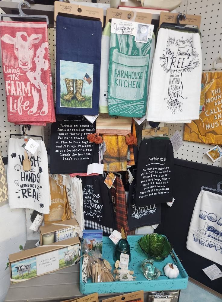 Display of farmhouse-themed kitchen towels with various sayings and designs, a tray of decorative items.