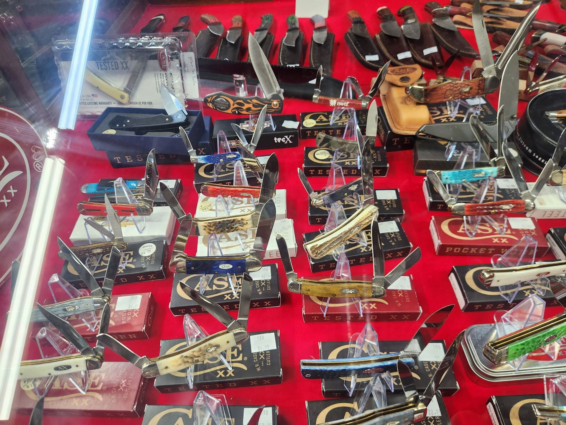 Display case of various folding knives, arranged on a red surface, with open blades and boxes.