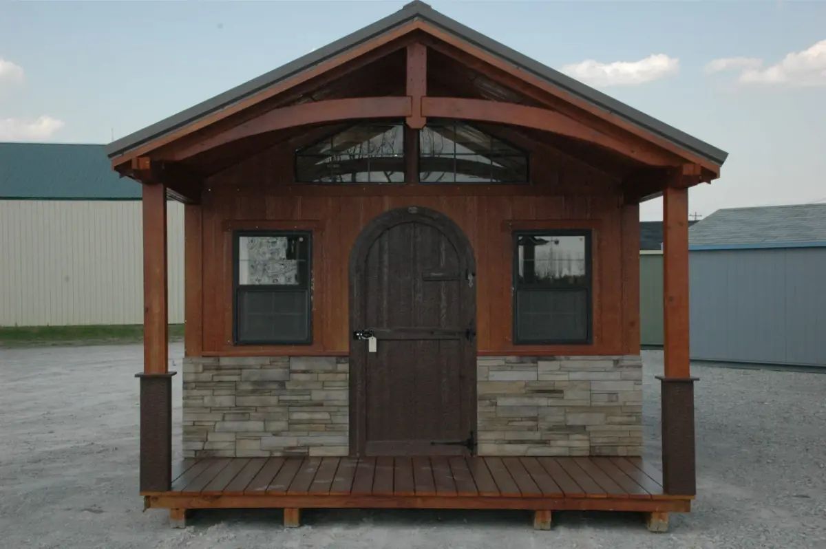 Small wooden shed with stone veneer and porch. Brown wood with a gothic door, two windows, and dark roof.