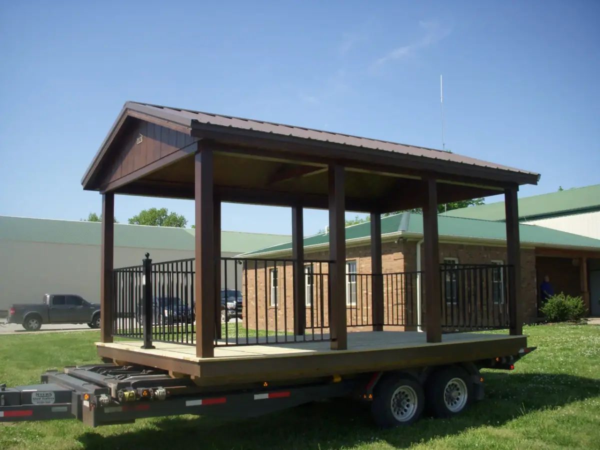Brown gazebo structure on a trailer, set outdoors on grass. Metal roof, black railing, and wooden posts.