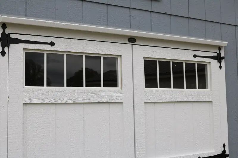 White carriage-style garage doors with rectangular windows, black hinges, and an overhanging white trim.