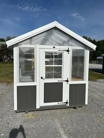 Small greenhouse with white trim and gray panels on a gravel lot under a blue sky.