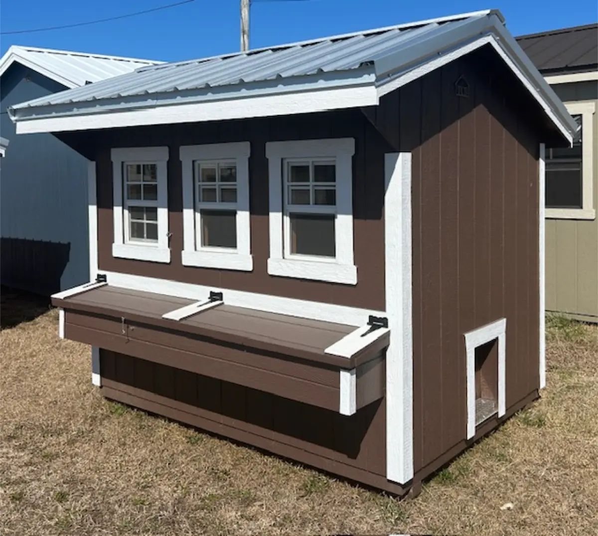 Brown chicken coop with three windows, a nesting box, and a doorway, on grass.