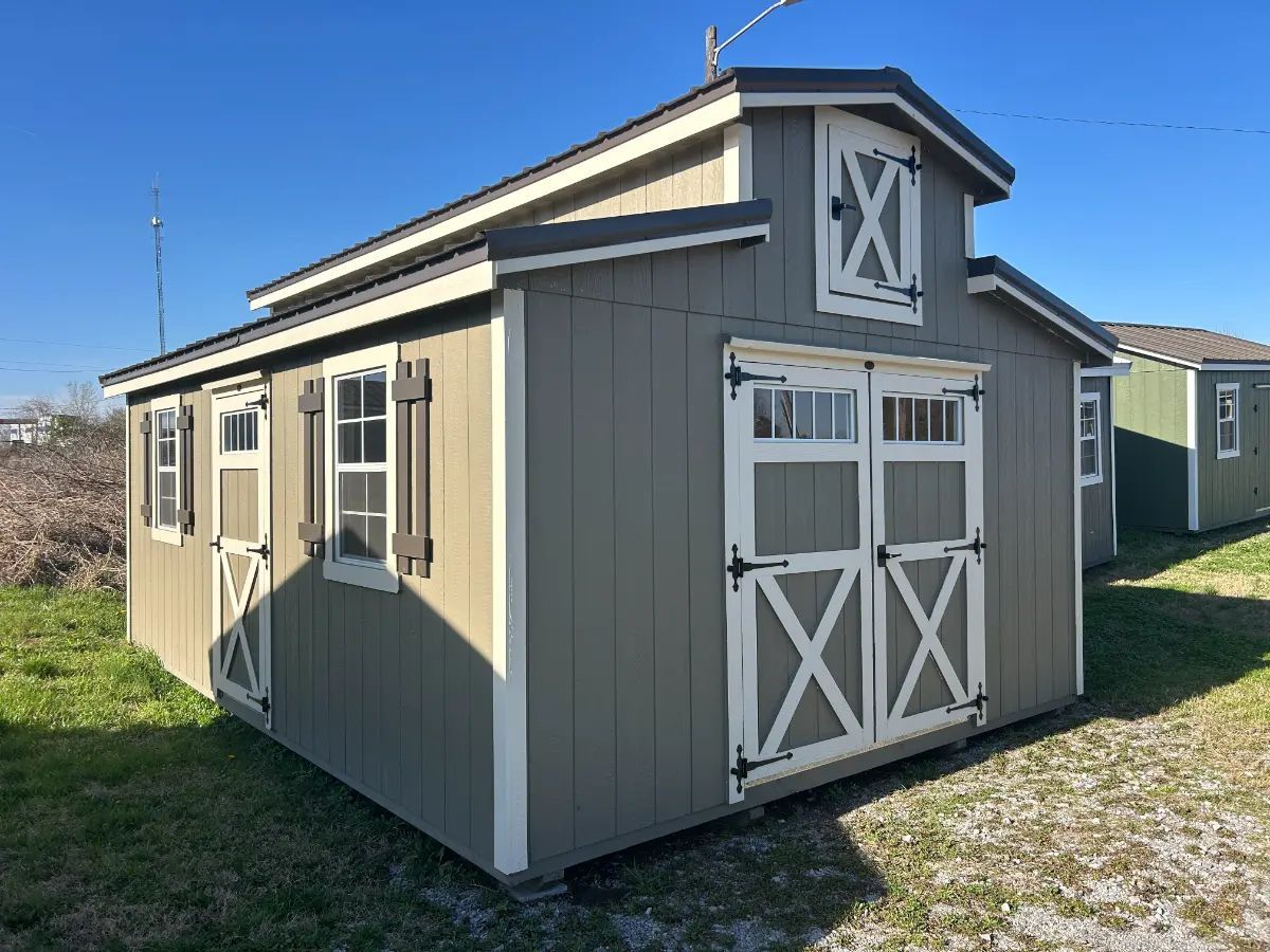 A two-story gray shed with white trim, double doors, and windows on green grass under a blue sky.