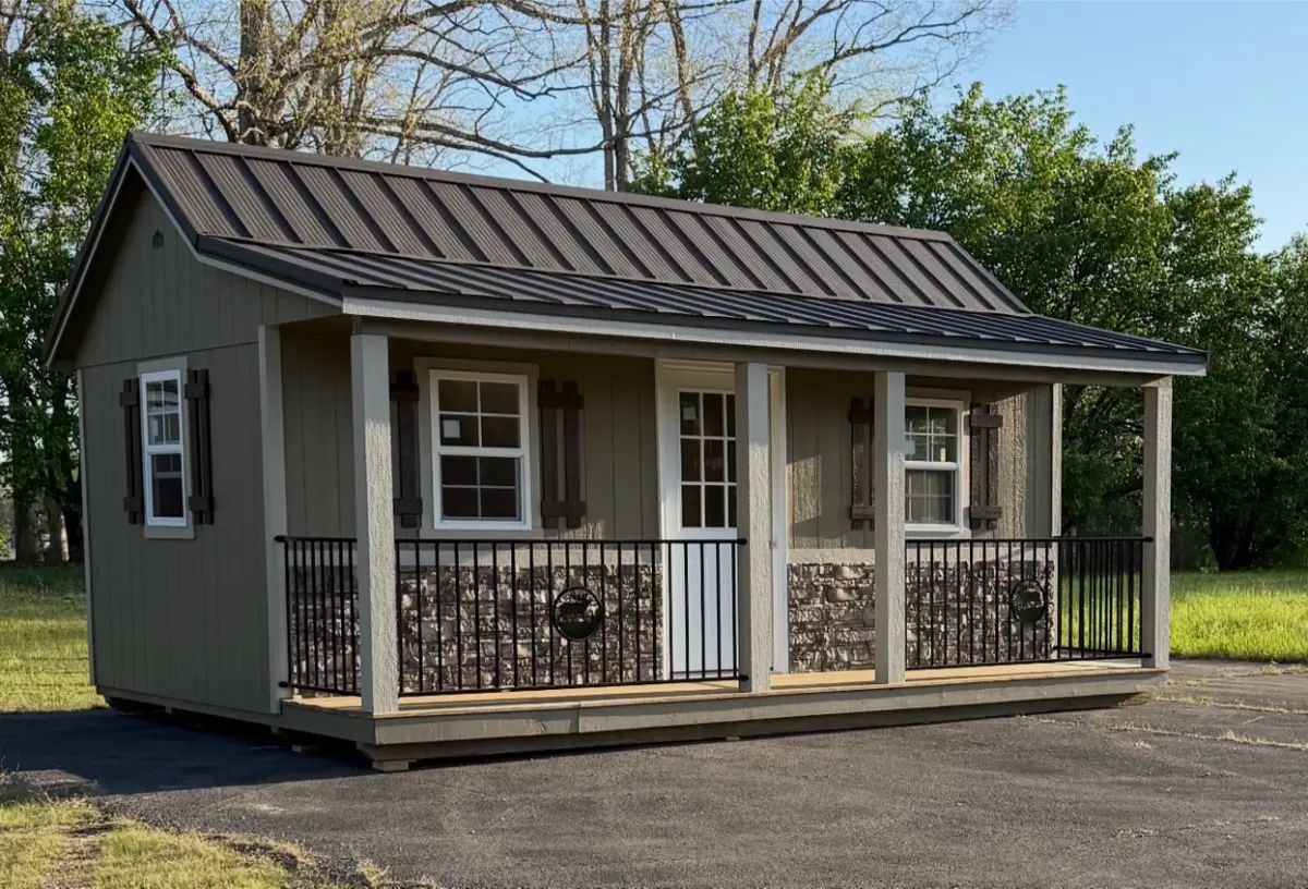 Small, tan cabin with black roof, porch, and decorative stone accents. White windows and door, black railing.