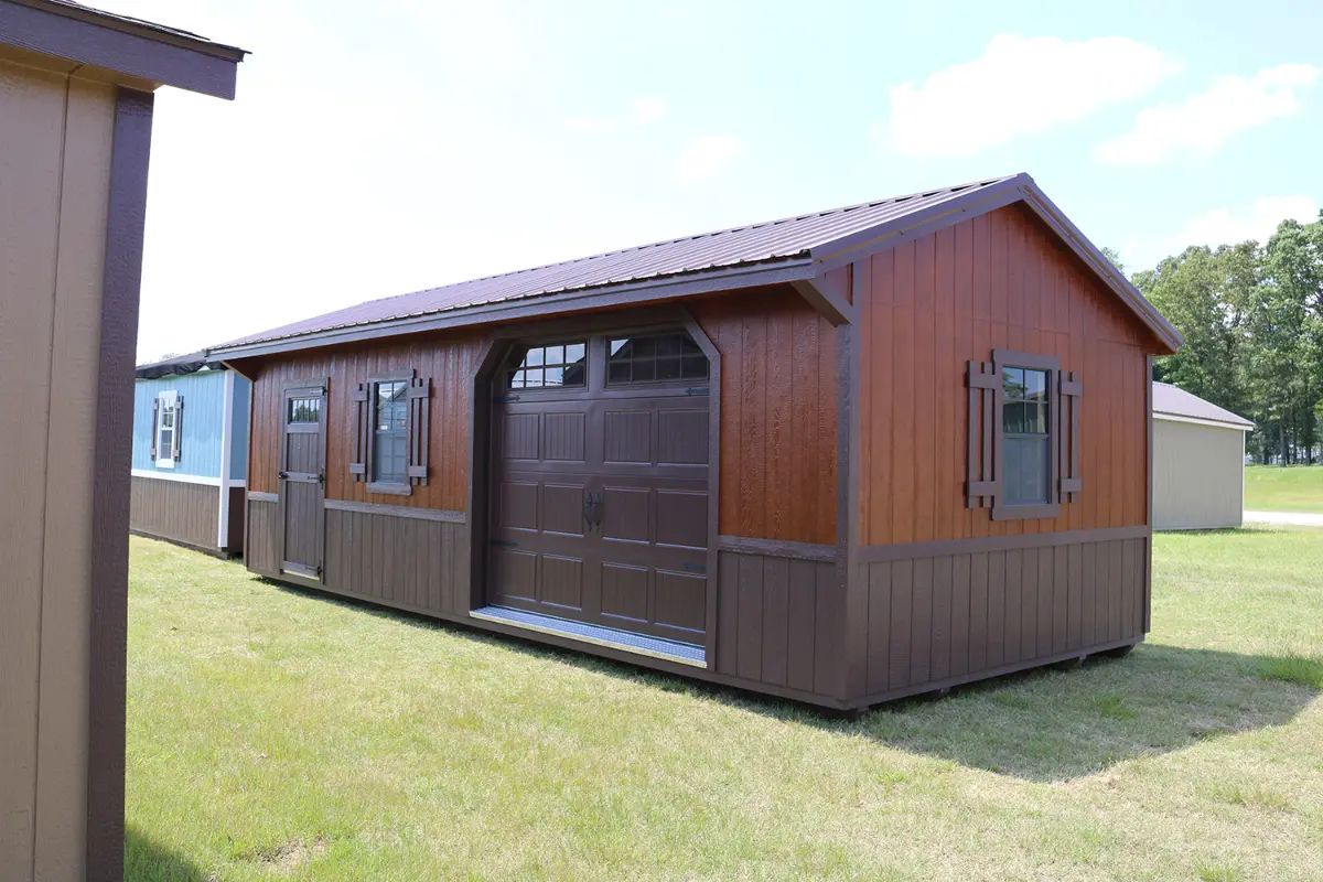 Brown and tan shed with garage door, windows, and a metal roof, set on green grass.