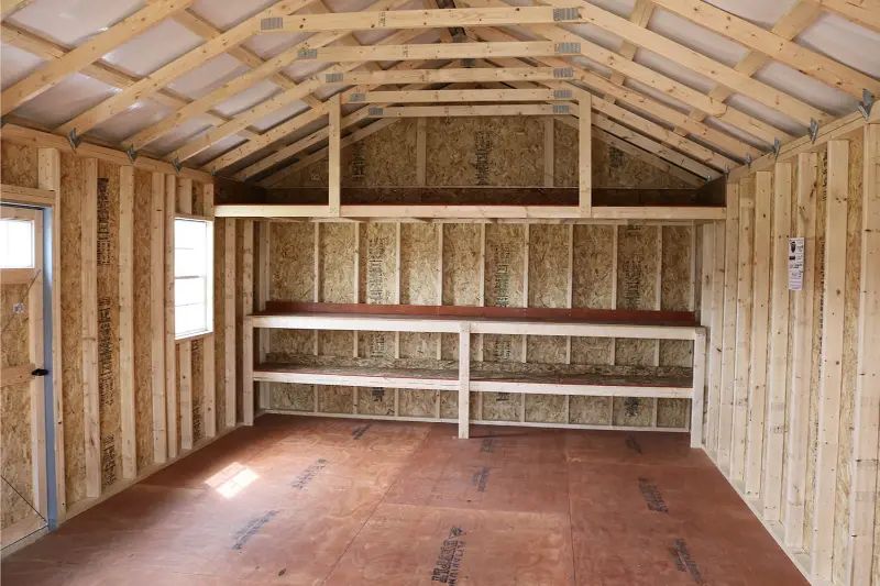 Interior view of a wooden shed with a workbench and loft storage, lit by a window and door.