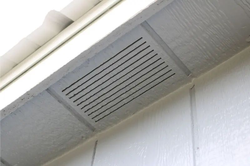 Ventilation grill on a house's exterior, positioned under the eaves. White siding and soffit with black slats.