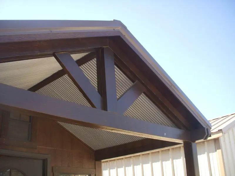 Brown wooden porch roof with a truss design, metal corrugated ceiling, and a blue sky background.