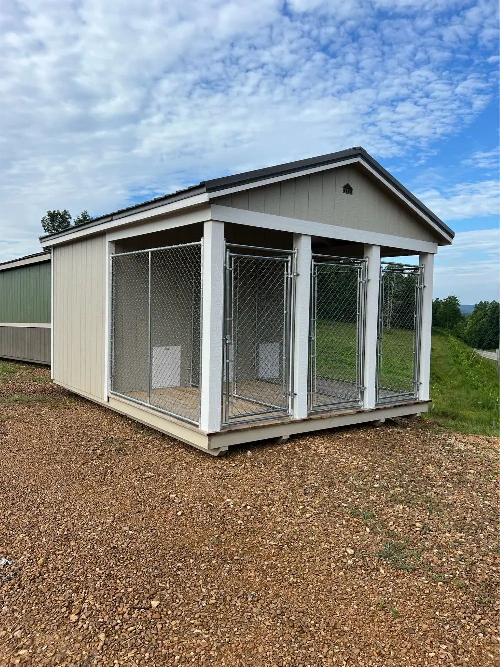 A beige dog kennel with three wire cages, under a roof, set on gravel. Overcast sky in the background.