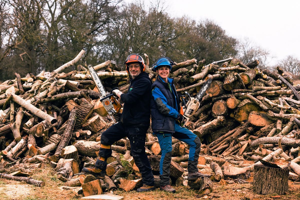 Two men are standing in front of a pile of logs holding chainsaws.