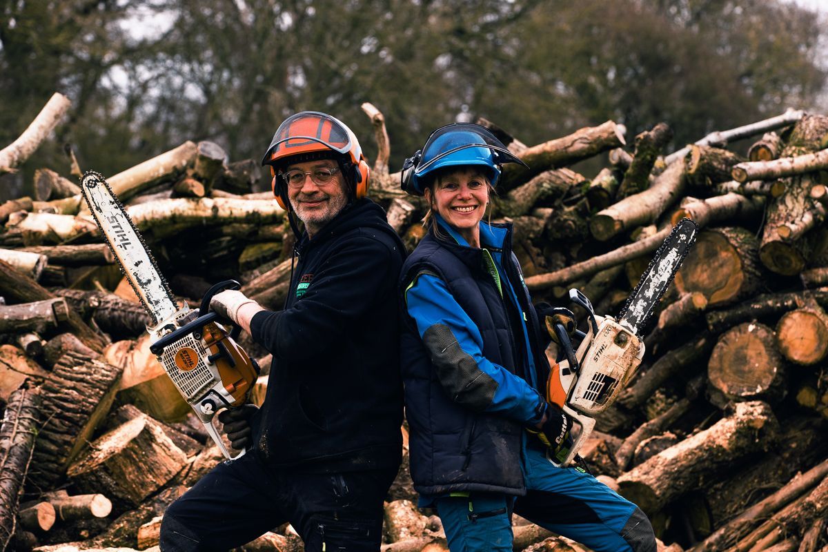 A man and a woman are holding chainsaws in front of a pile of logs.