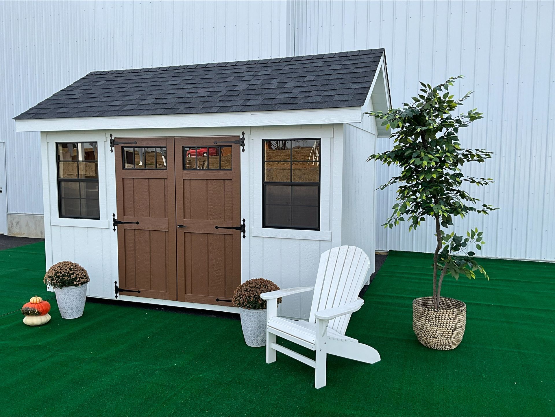 White shed with brown double doors, 2 black windows, and dark asphalt roofing.