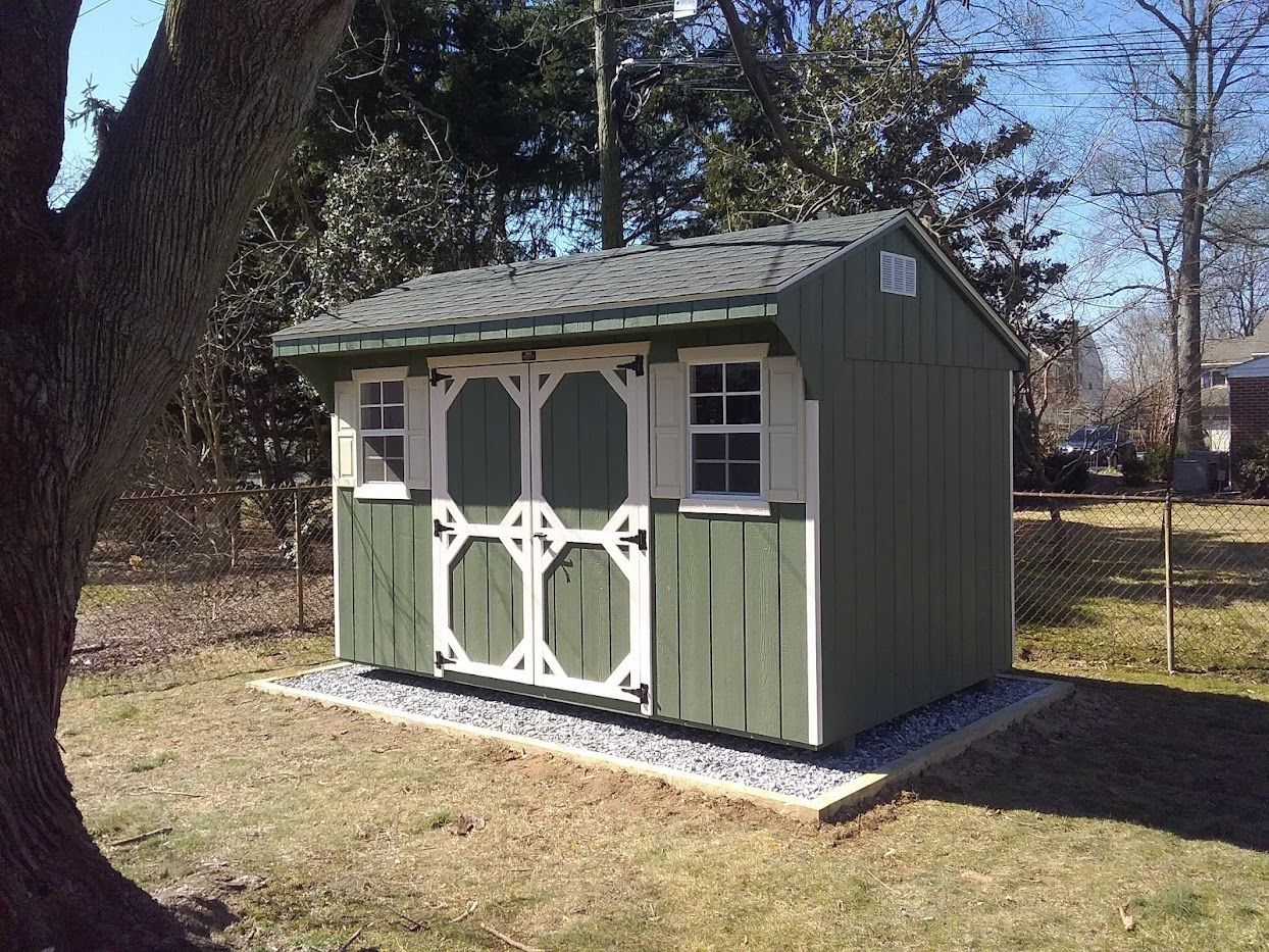 Quaker Shed with green wood siding, white trim, a set of double doors, white windows with white shutters, and a gray asphalt roof on a stone pad foundation in a backyard.