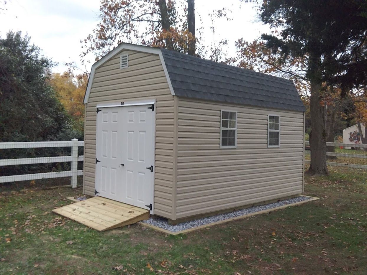 Tan vinyl shed with white double doors, a wood ramp, 2 white windows, and a gray asphalt roof.