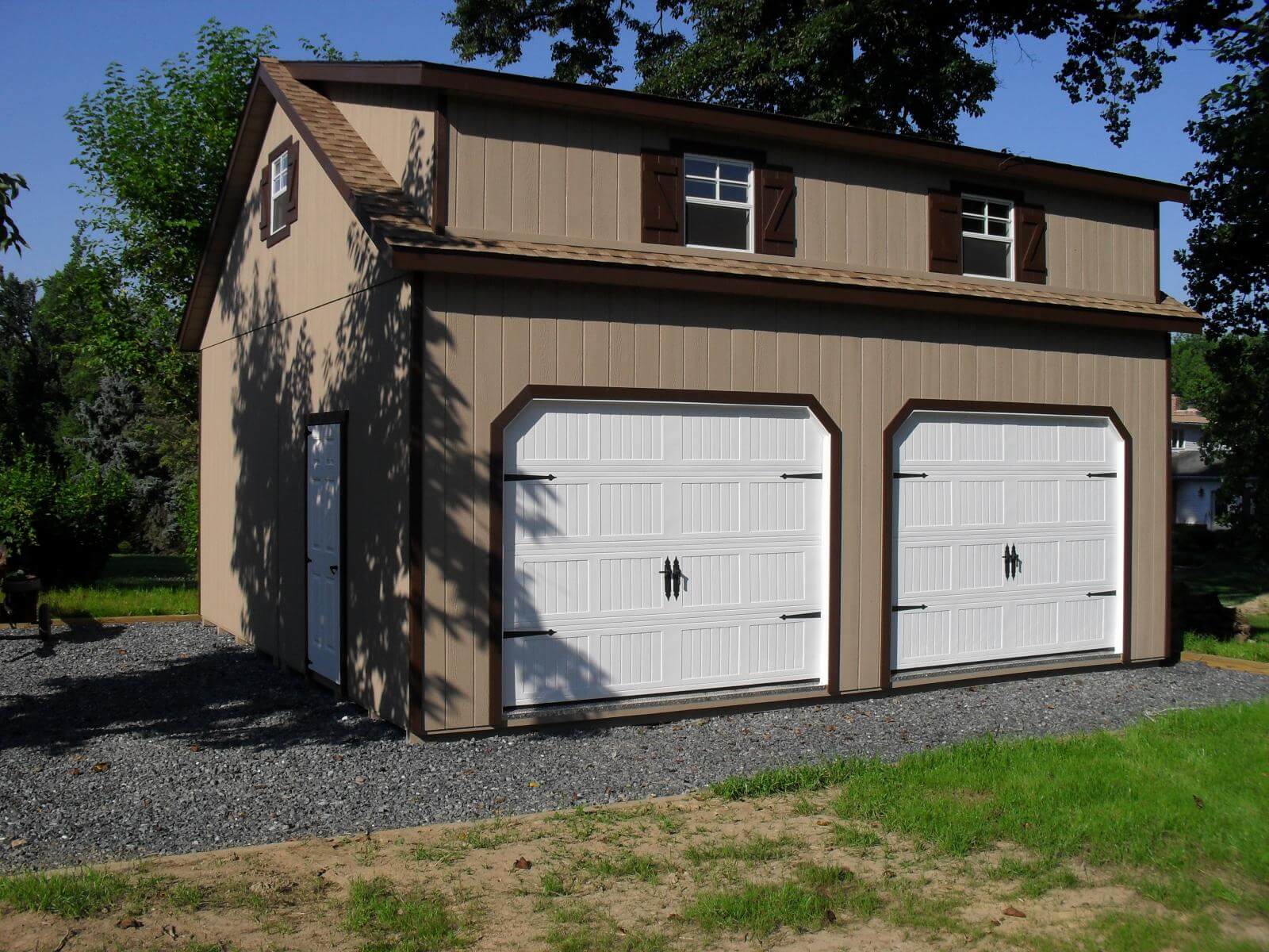 Tan two-story two-car garage with 2 white garage doors, dark brown trim, brown roofing, and windows with dark brown shutters