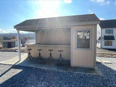 Tan pool shed with gray-tan trim, a covered counter with 4 bar stools, brown asphalt roofing, and a single black window.