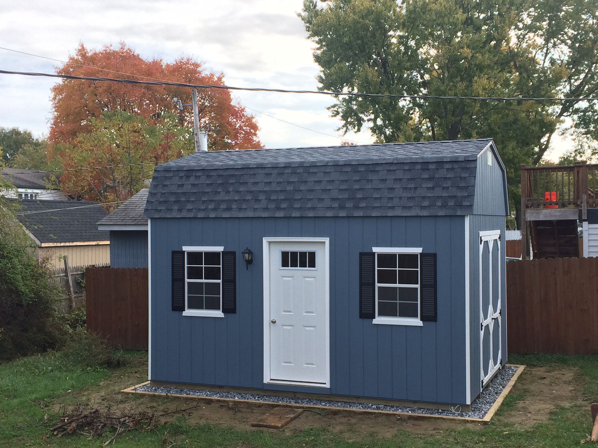 Blue shed with white trim, 2 white windows with black shutters, a white single-entry door with transom windows, and a gray asphalt roof.