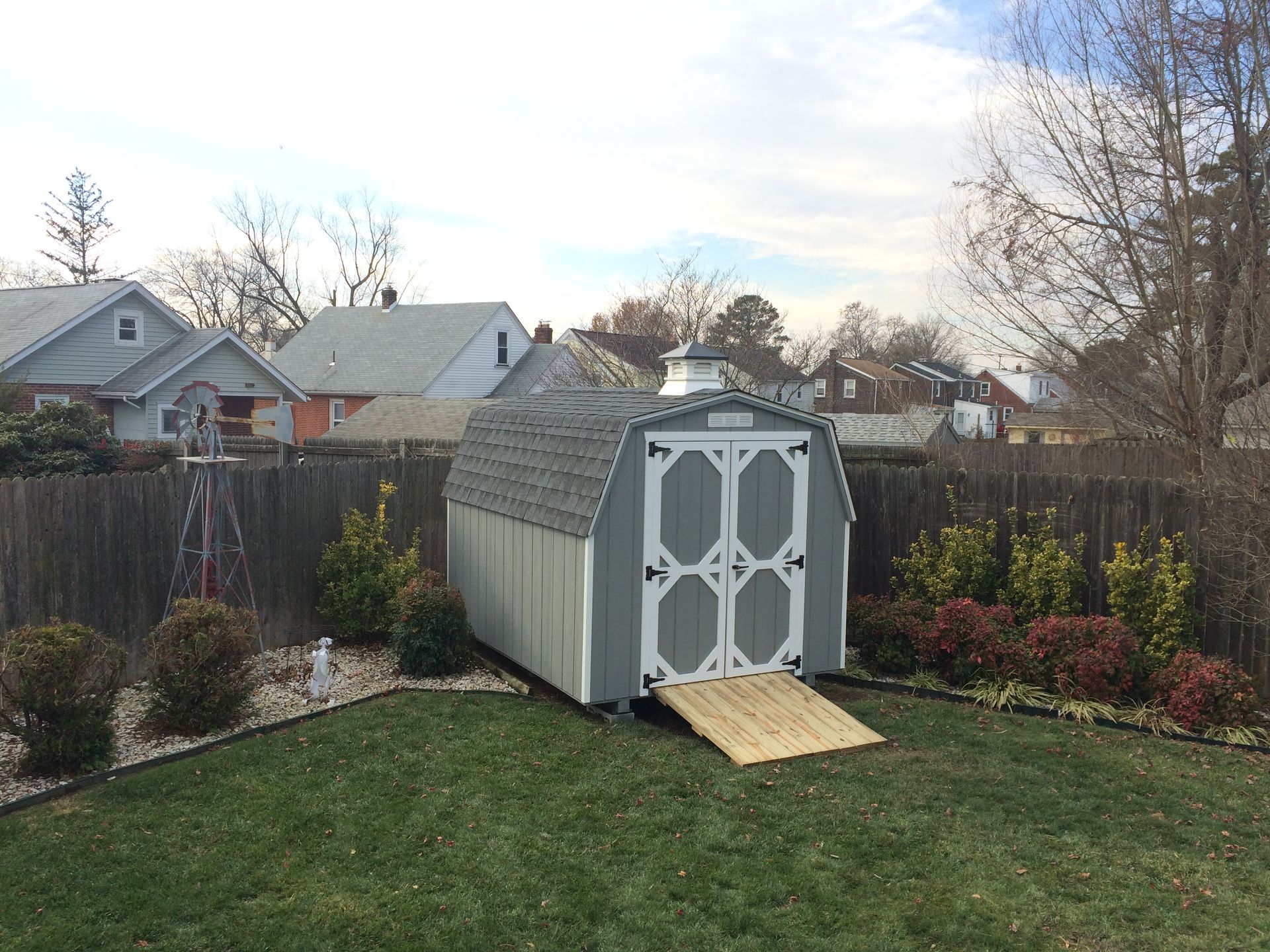 Mini Barn Shed with gray wood siding, white trim, a set of double doors, a wood ramp, a white gable vent, a gray asphalt roof, and a white and gray cupola sits in the corner of a yard against a wood fence.