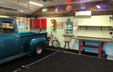 interior of a garage with a blue truck, workbench, carpeted floors, and wall art