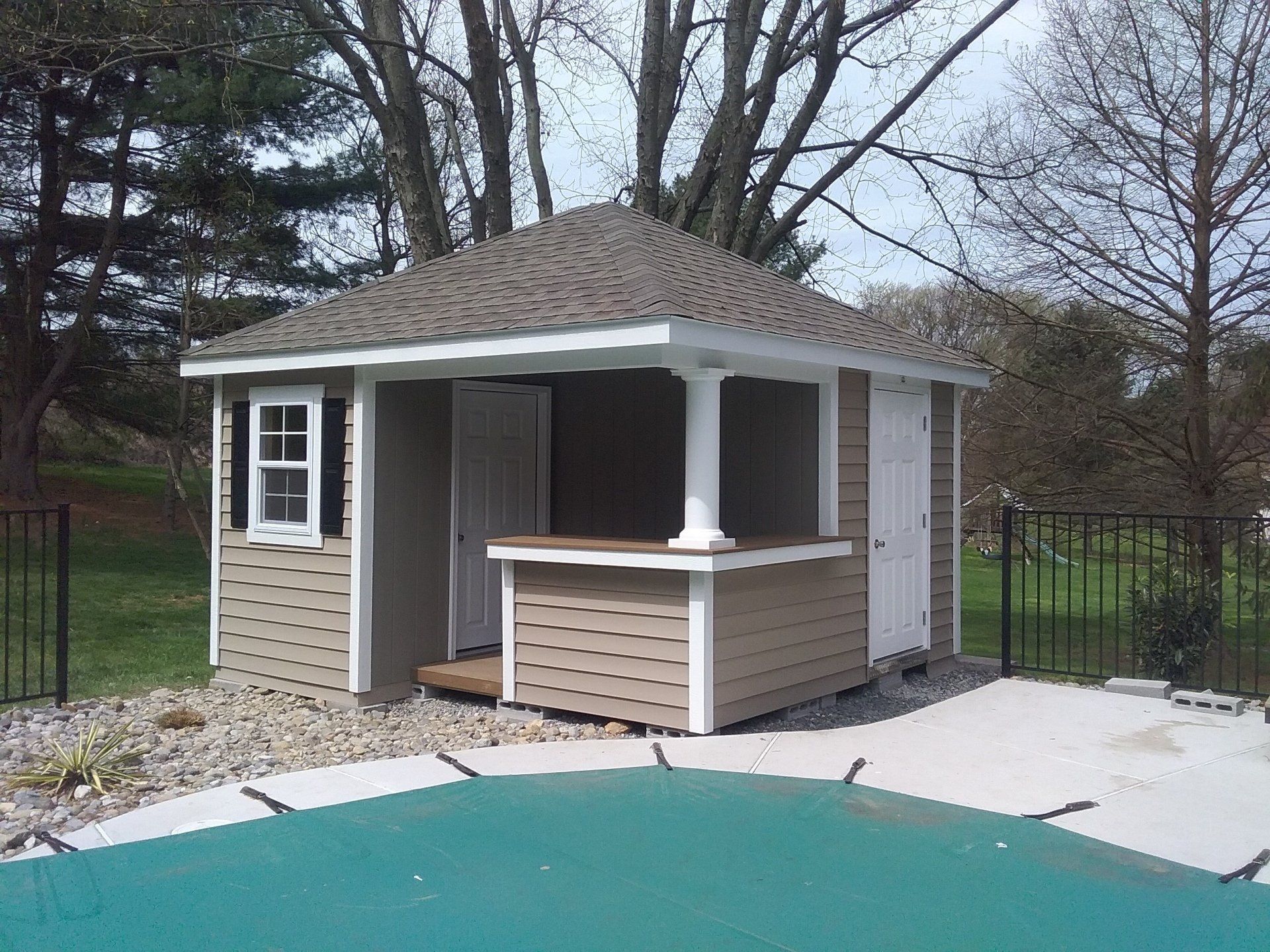 Pool house with tan siding, white trim, 2 white single doors, a window with black shutters, and a brown roof
