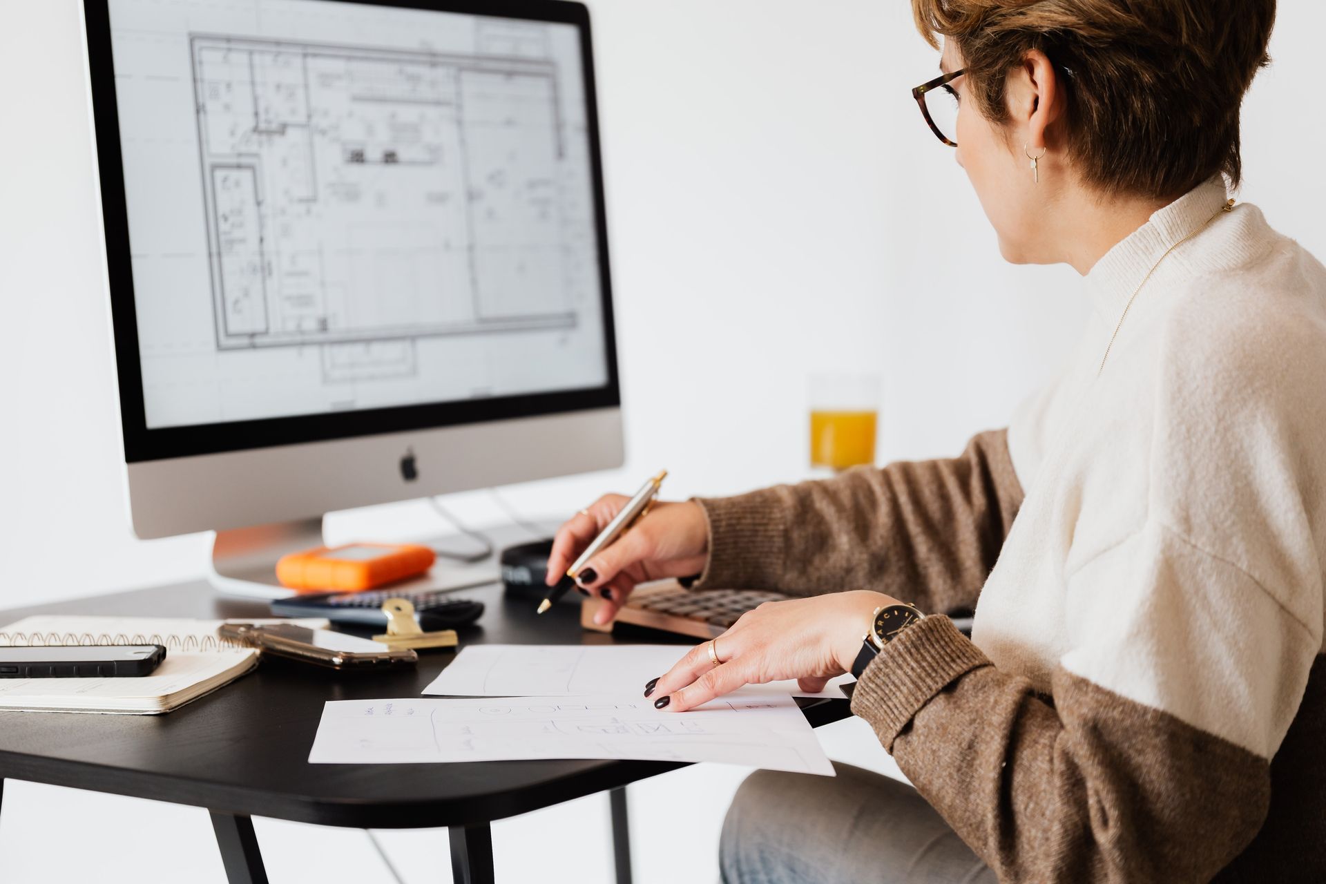 Person in white and brown sweater writes on paper while looking at a blueprint on their monitor.