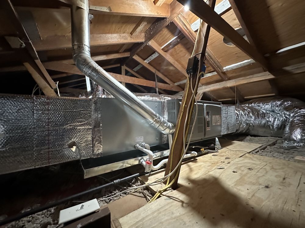 HVAC unit in a dusty attic with ductwork and wooden beams overhead.