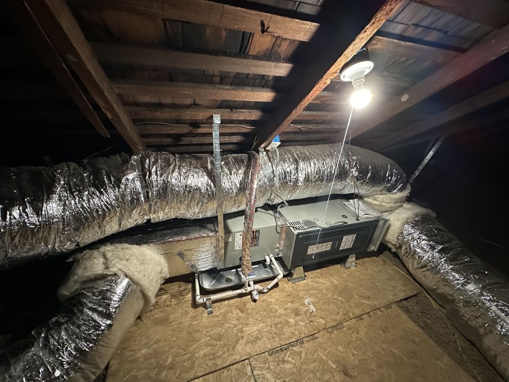 HVAC unit in an attic with insulated ductwork, wooden beams, and a work light overhead.