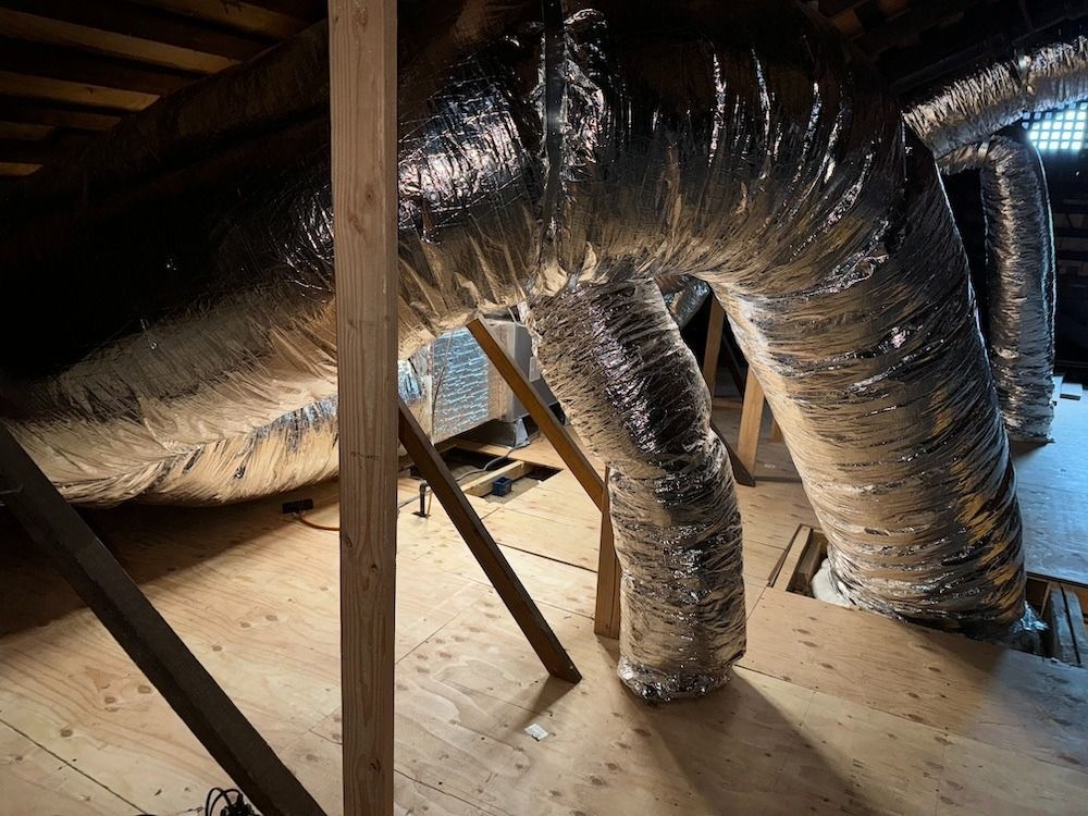 Flexible metallic HVAC ductwork snaking through an attic with exposed wooden supports.