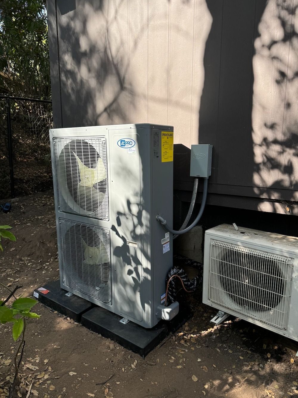 Two air conditioning units outside a building. One large unit with two fans sits on a black pad.