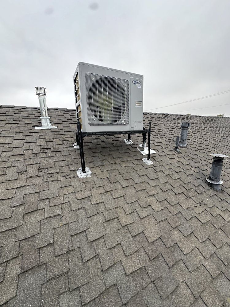 HVAC unit on a shingled roof, supported by black metal legs, against a cloudy sky.