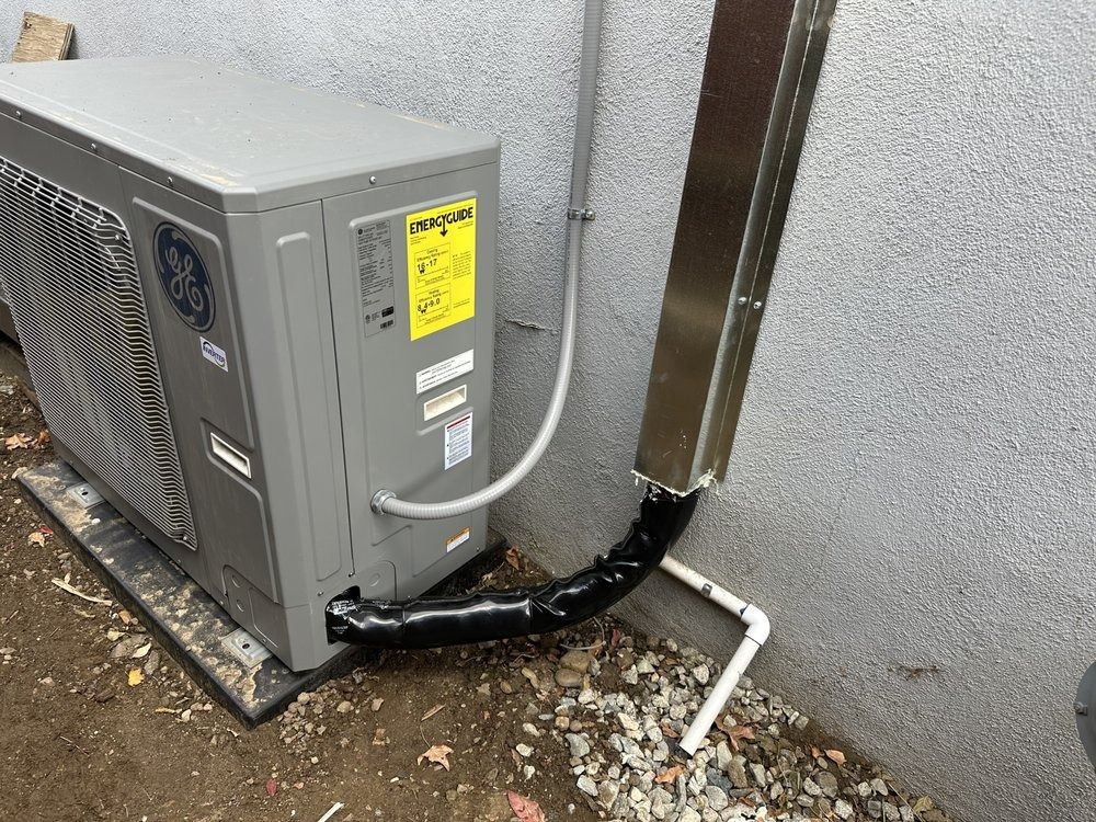 Gray heat pump unit next to a wall with a silver pipe.