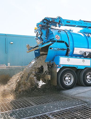 Blue vacuum truck expelling waste onto a grate near a blue building.