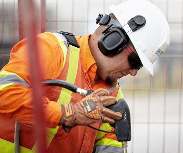 Construction worker wearing safety gear operates a tool near a fence; bright orange vest, white hard hat.