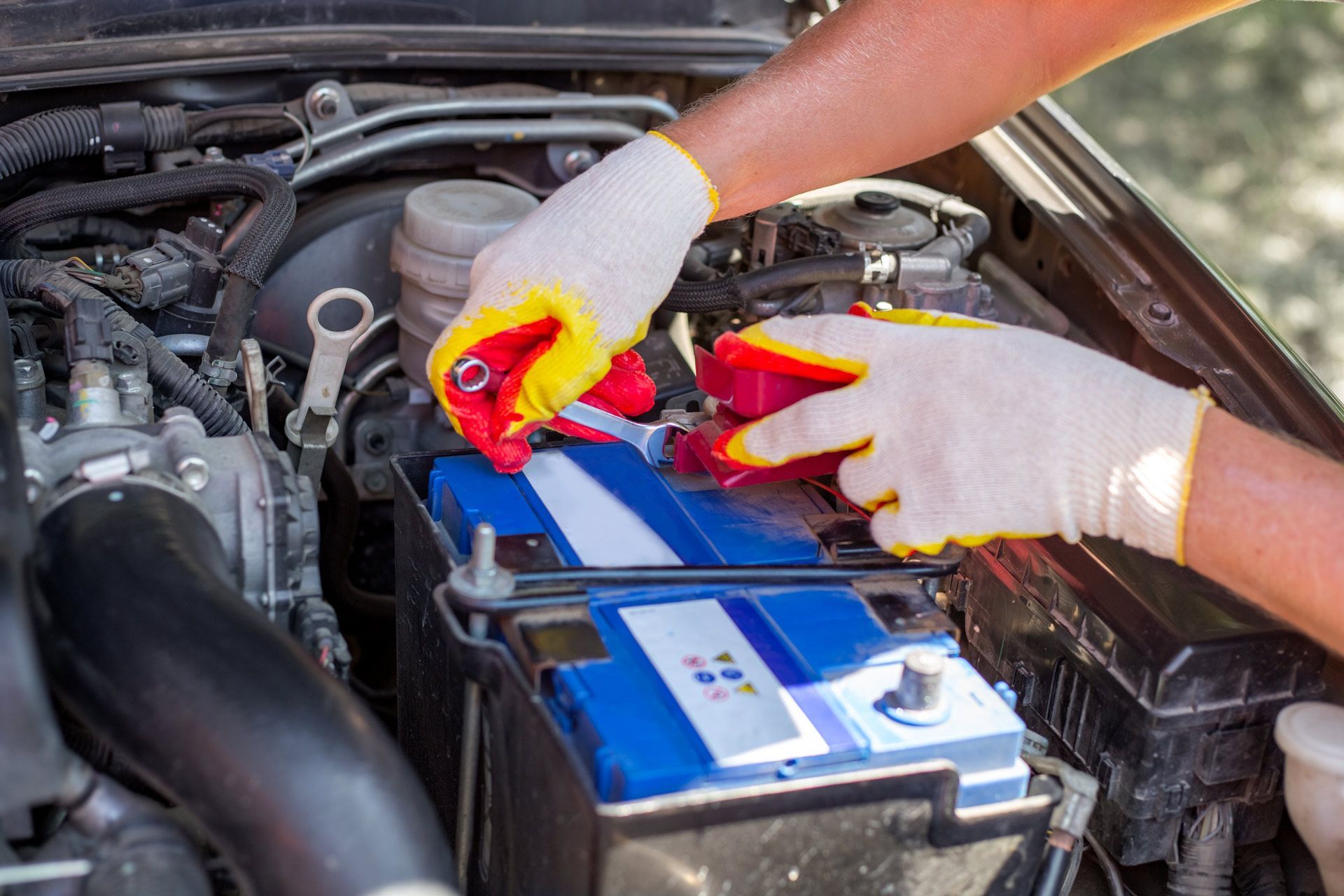 Hands in gloves working on a car battery with tools, inside the engine compartment.