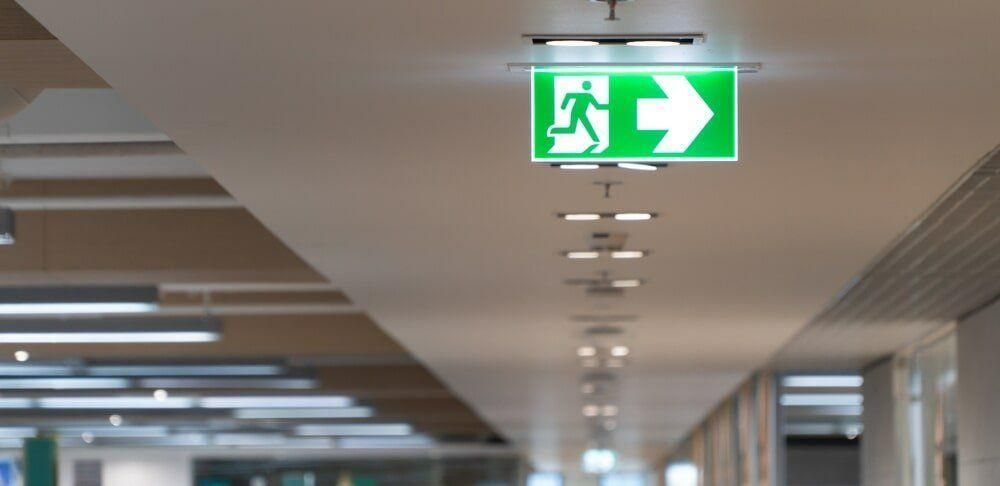 A Green Exit Sign Is Hanging From The Ceiling Of A Building — DG Electrical & Fire In Kiama, NSW