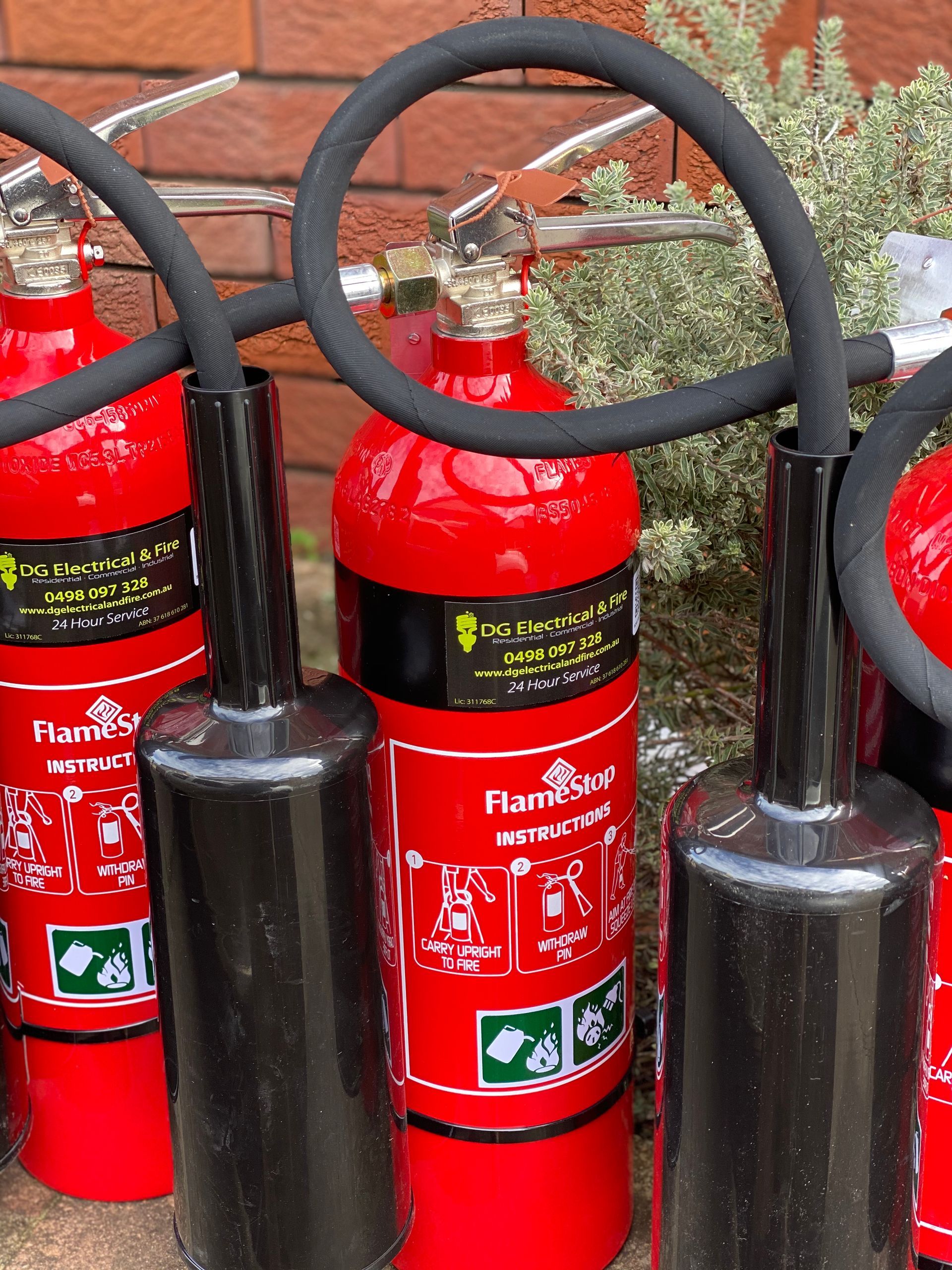 Red fire extinguishers with black hoses and nozzles lined up outdoors. — DG Electrical & Fire In Kiama, NSW