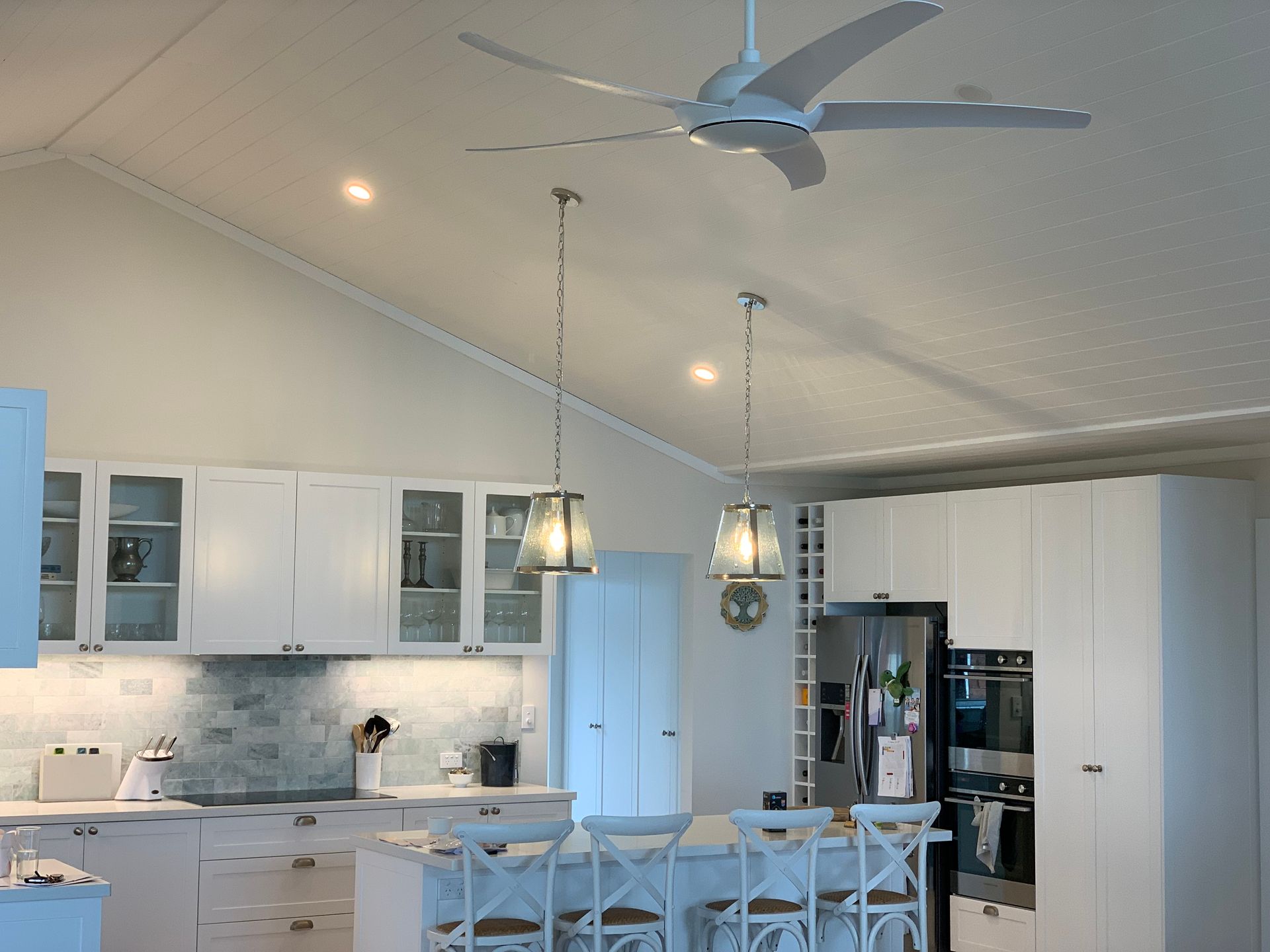 White kitchen with vaulted ceiling, pendant lights, island with stools, and ceiling fan. — DG Electrical & Fire In Kiama, NSW