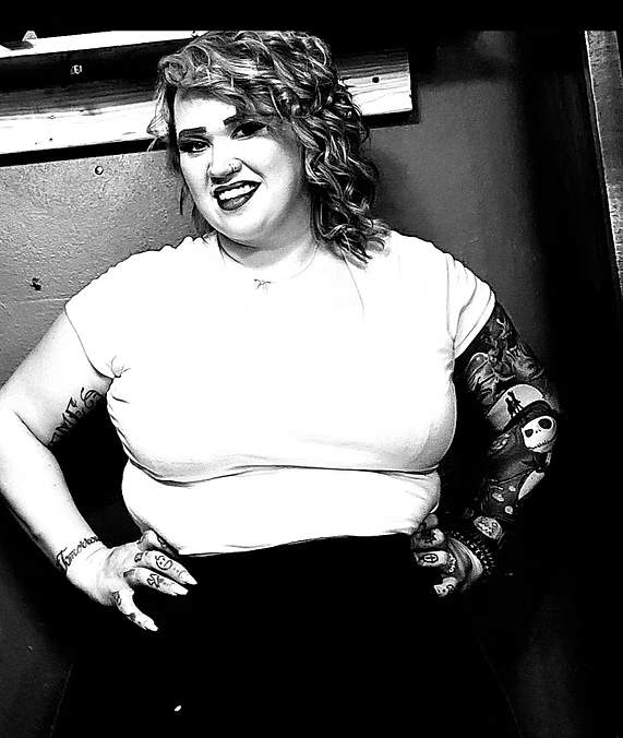 Woman in white shirt and dark skirt with tattoos, hands on hips, smiling.