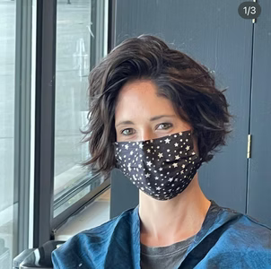 Woman with short dark hair, wearing a star-patterned face mask, near a window.