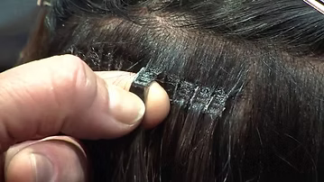 Hand holding a small, black, rectangular hair extension attached to a person's dark hair.