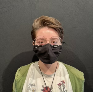 Person wearing glasses and a black face mask, posing against a dark background.
