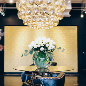 A round glass table with a vase of white roses sits under a large chandelier.