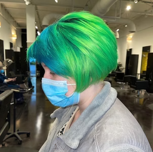 Woman with green and blue hair, wearing a mask, inside a hair salon.