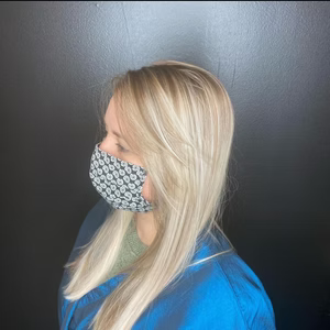 Woman with blonde hair wearing a patterned face mask and blue jacket against a black background.