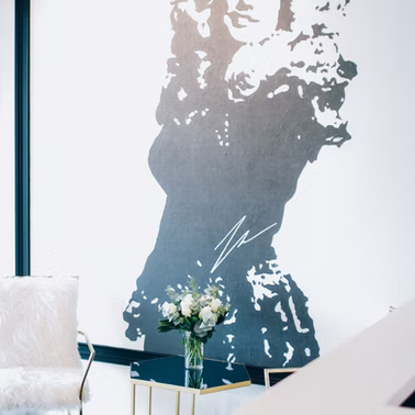 Black and white mural of a person. A small bouquet sits on a gold table. White furry chair to the left.